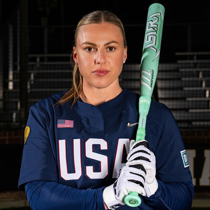 A focused softball player in a navy USA jersey holds the 2025 Marucci ASURA (-9) Fastpitch Softball Bat (MFPAS9 DEMO) with Gap Sync barrel technology, wearing white batting gloves and her blonde hair pulled back, standing before stadium seats.