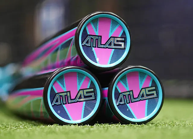 Three baseball bats stacked in a pyramid, with ATLAS logos visible on the end caps. The bats feature a vibrant blue, pink, and black geometric design. They rest on green artificial turf.