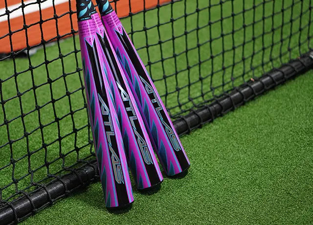 Three purple and blue baseball bats with ATLAS branding lean against a black net on artificial green turf near an orange boundary line.
