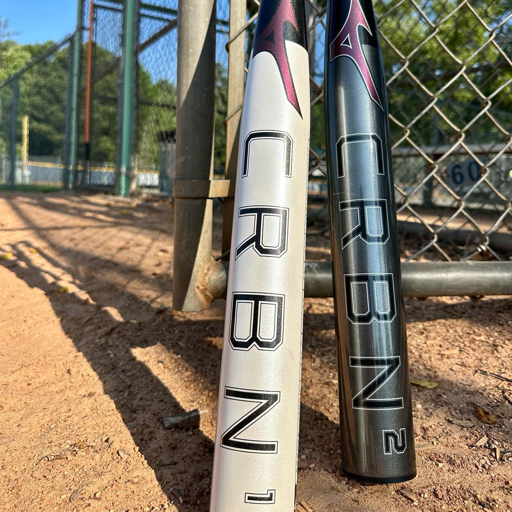 Two Mizuno CRBN baseball bats, labeled CRBN 1 and CRBN 2, are standing upright on a dirt field near a chain-link fence, with a gate and trees visible in the background.