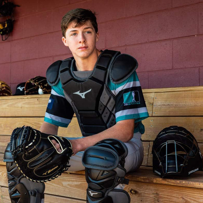 A teenage baseball catcher in full gear sits on a wooden bench in a dugout, looking ahead. His helmet rests on the bench beside him, and several gloves hang on the wall in the background.