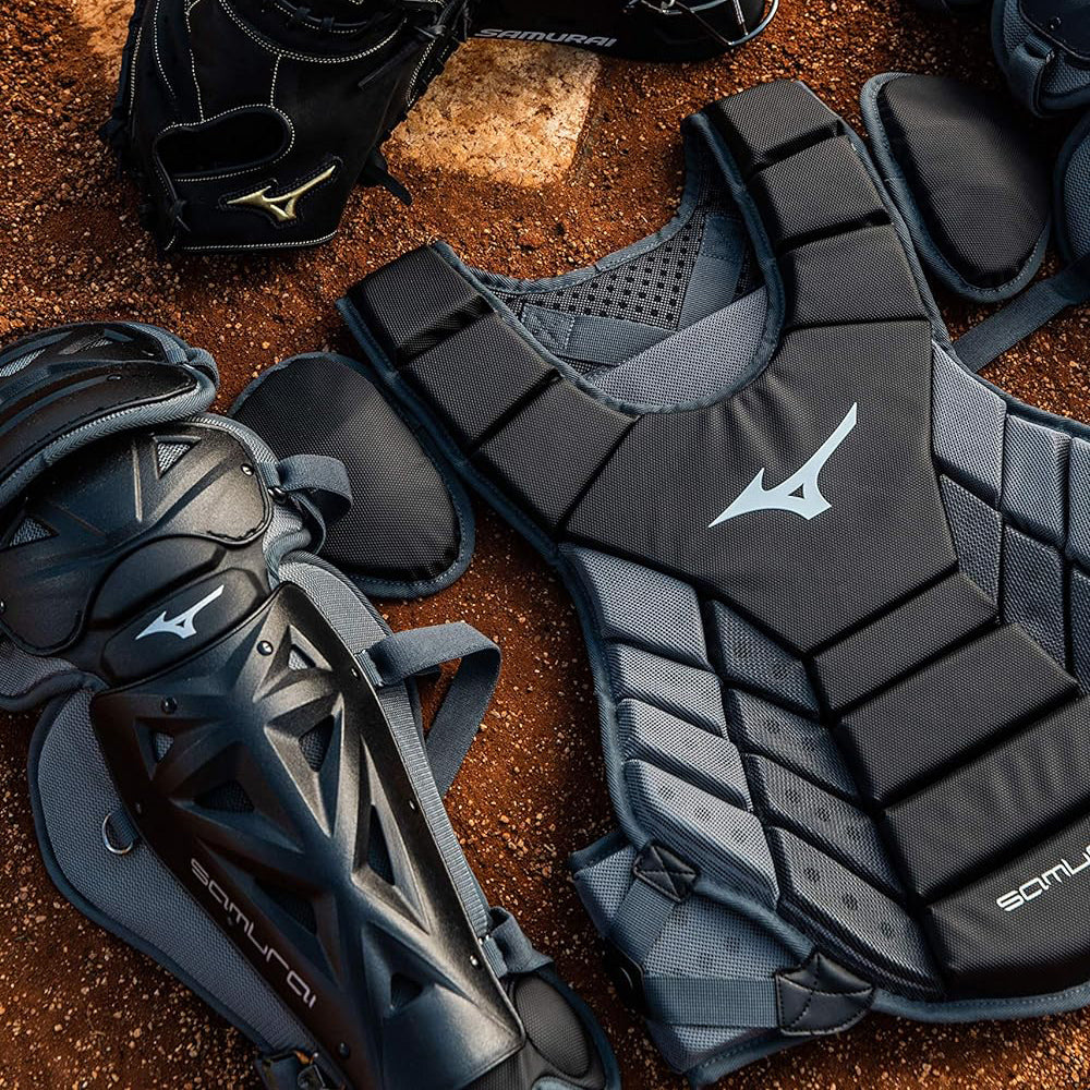 A set of black and gray baseball catchers gear, including a chest protector, shin guard, and glove, is laid out on a dirt surface near home plate.