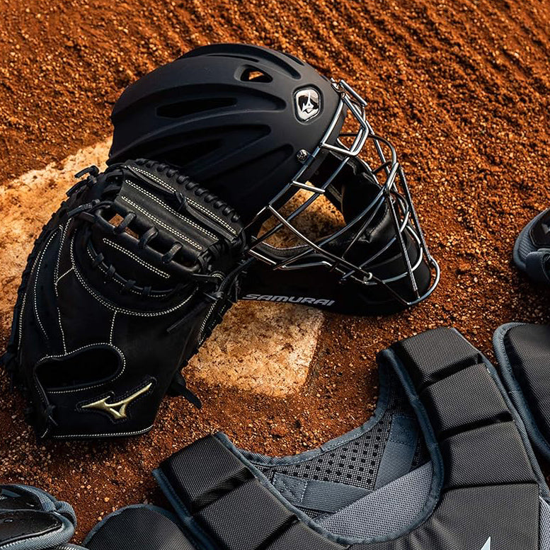 A black baseball catchers helmet and glove rest on a pitchers mound, surrounded by protective gear and chest padding on the reddish-brown dirt field.