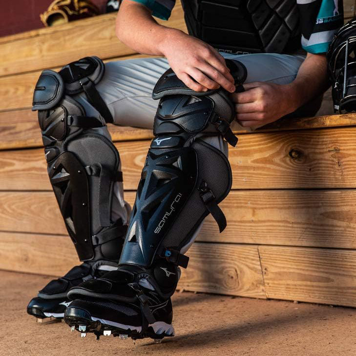 A baseball catcher sits on a wooden bench, wearing protective gear including shin guards, chest protector, and cleats, preparing for a game.