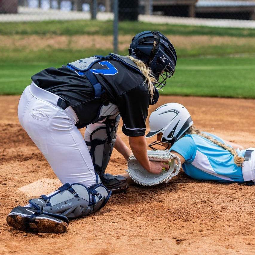 A softball catcher in black gear tags a sliding runner in light blue and white at home plate during a game, both players focused on the play on a dirt field.