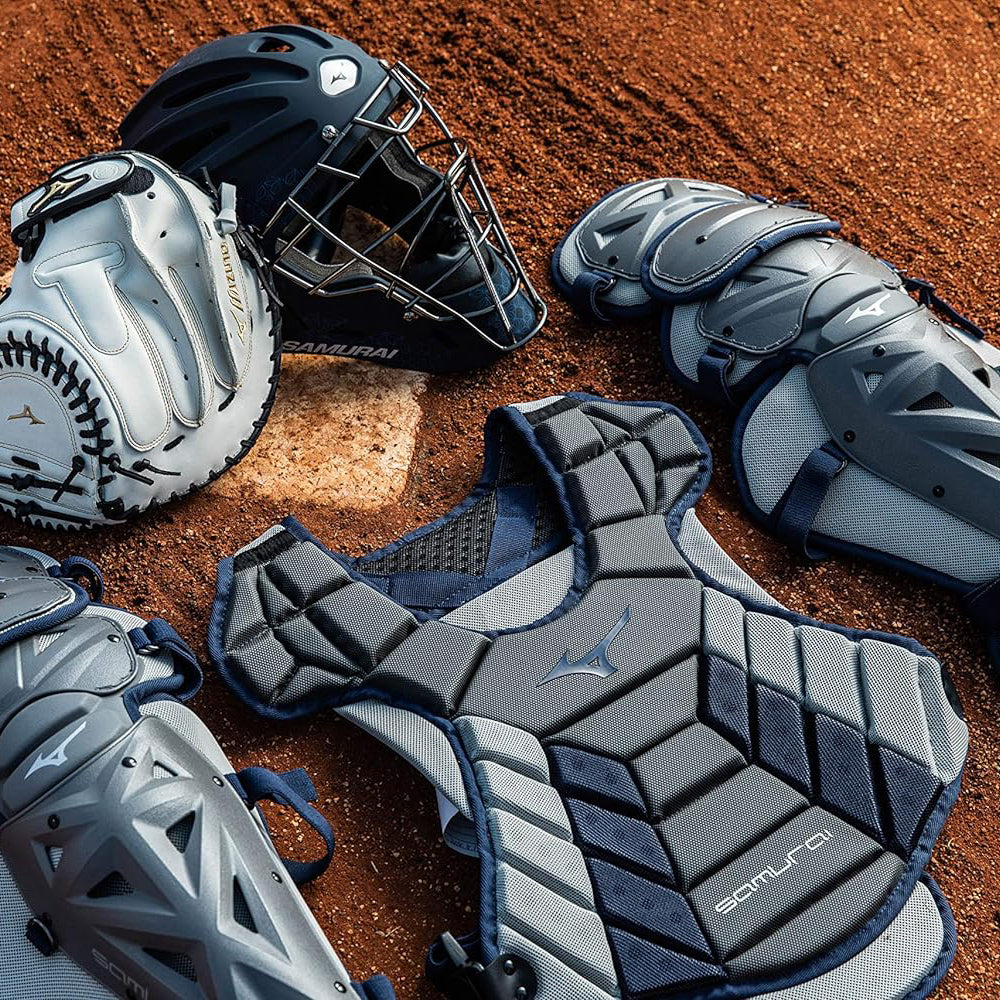 A set of gray and blue baseball catcher’s gear, including a helmet, chest protector, leg guards, and a glove, is arranged on a dirt field near home plate.