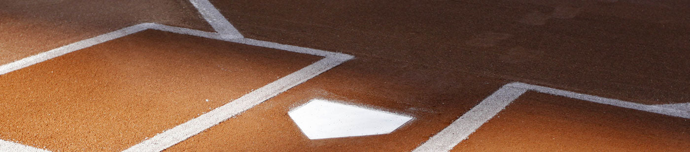 A baseball home plate on a dirt field with a puddle of water reflecting the sky, surrounded by white chalk baselines.