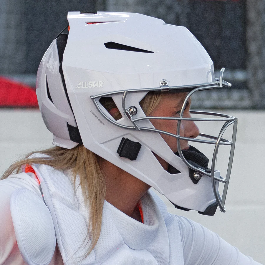 A softball catcher in an All Star PHX Paige Halstead MVP Pro Hockey Style Catcher's Helmet (MVP5-PHX), white, faces right during a game, with the All Star brand visible on the helmet.