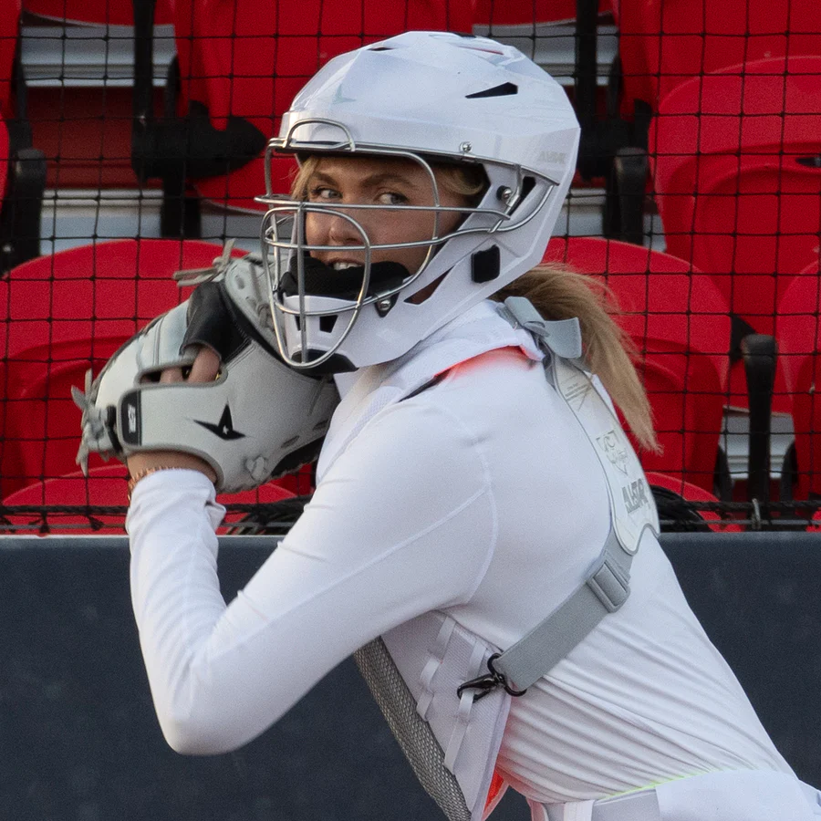 A softball catcher in an All Star PHX Paige Halstead MVP Pro Hockey Style Catcher's Helmet (MVP5-PHX) holds a mitt up, ready to throw, with red stadium seats and a black net in the background—ensuring top protection on the field.