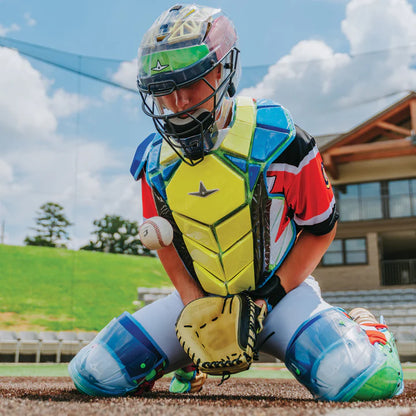 A catcher wearing the All Star MVP Pro Catcher's Kit CKCC-5 by All Star kneels on the field, stopping a ball with his chest protector. Bleachers and a building appear in the background under a partly cloudy sky.