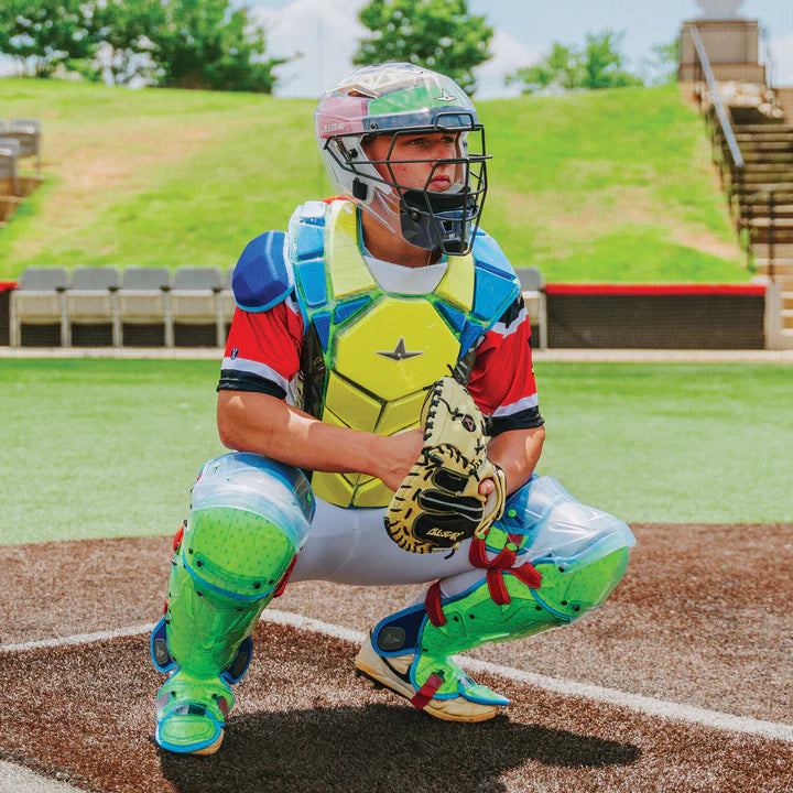 Wearing the All Star MVP Pro Catcher's Kit (CKCC-5), a catcher crouches behind home plate on an outdoor field, ready to receive a pitch. Bleachers and green trees are visible in the background under a sunny sky.