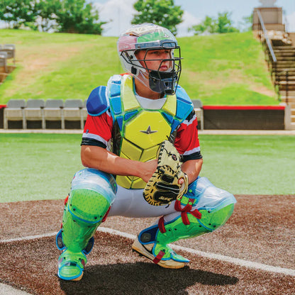 Wearing the All Star MVP Pro Catcher's Kit (CKCC-5), a catcher crouches behind home plate on an outdoor field, ready to receive a pitch. Bleachers and green trees are visible in the background under a sunny sky.