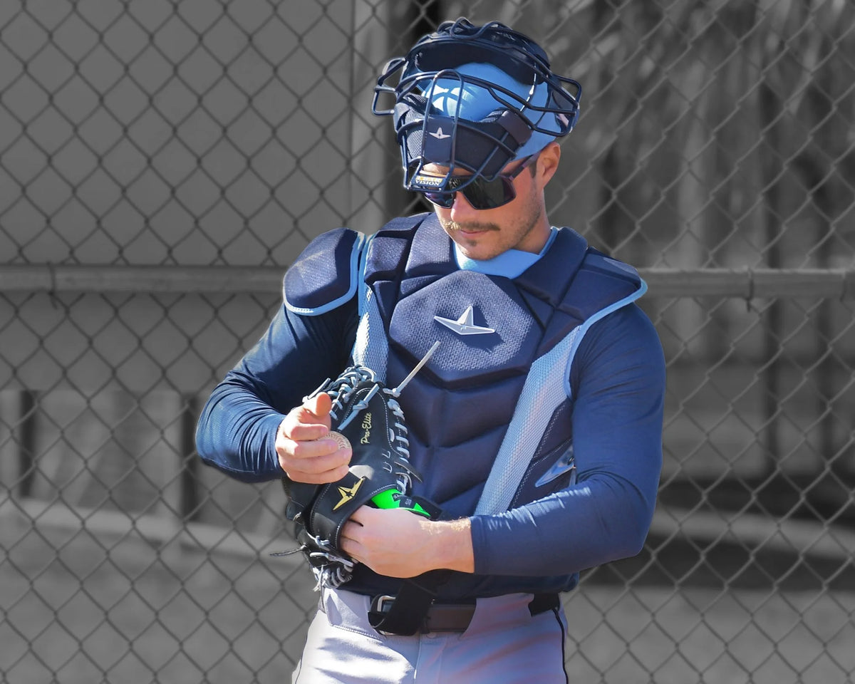 Wearing full gear, a baseball catcher adjusts his black All Star MVP-PRO Catcher’s Thumb Guard (CTG-MVP-LGE-LG) glove in front of a chain-link fence during daylight.