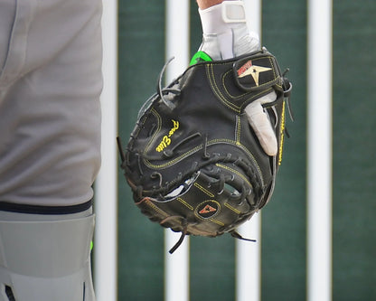 A person wearing a gray uniform and white glove holds an All Star MVP-PRO Catcher’s Thumb Guard (CTG-MVP-LGE-LG) by All Star, designed for superior thumb protection. Vertical white bars and a green background are visible behind them.