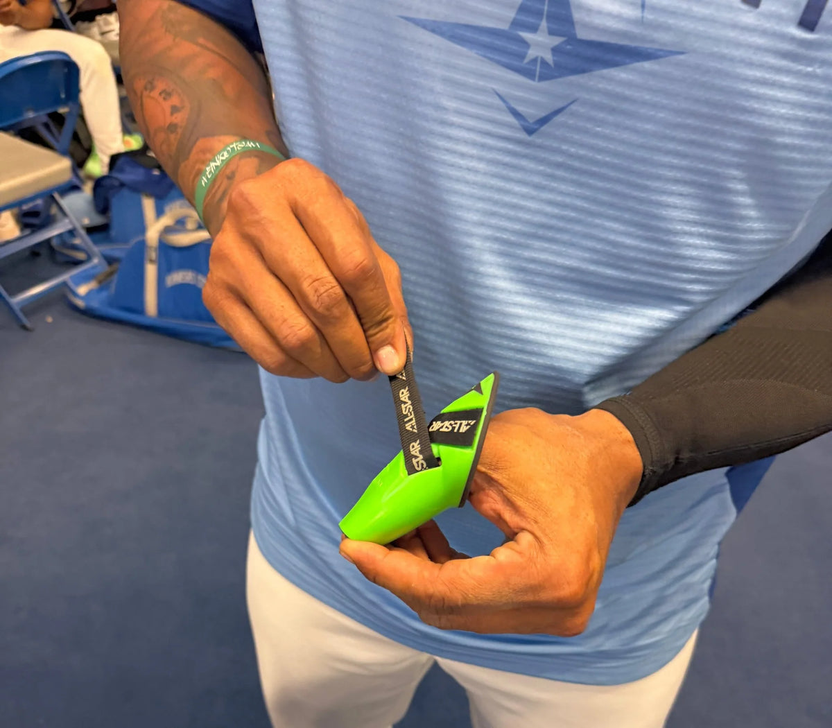 A person wearing a blue shirt opens a green packet labeled ASK with a black tear strip, their hands and torso visible as they demonstrate proper thumb protection using the All Star MVP-PRO Catcher’s Thumb Guard (CTG-MVP-LGE-LG) by All Star.