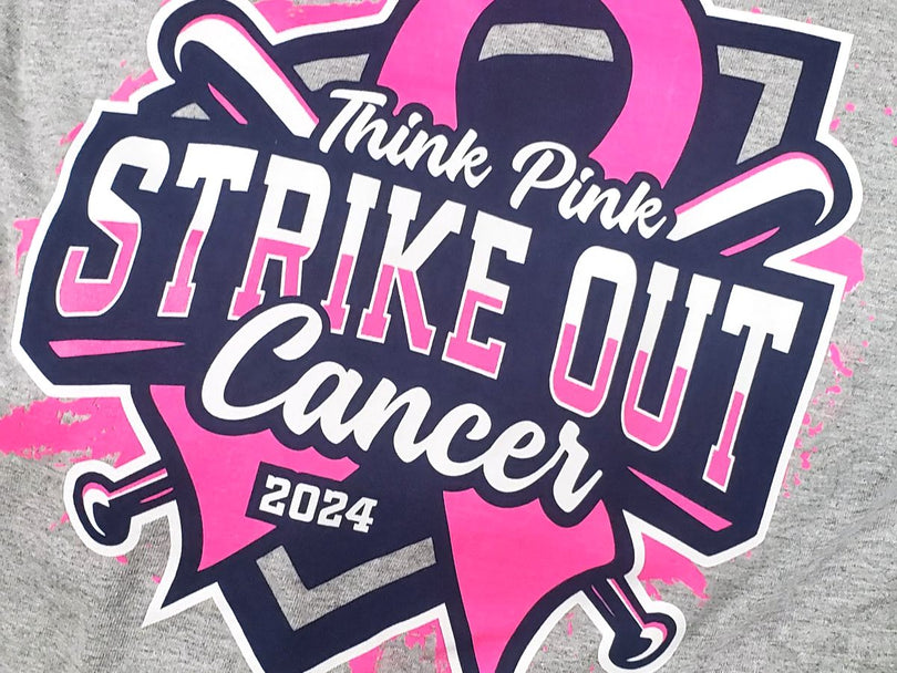Gray shirt with a bold design featuring the words “Think Pink Strike Out Cancer 2024” in pink, white, and black letters, with two crossed bats, a large pink ribbon, and pink paint splatter accents.