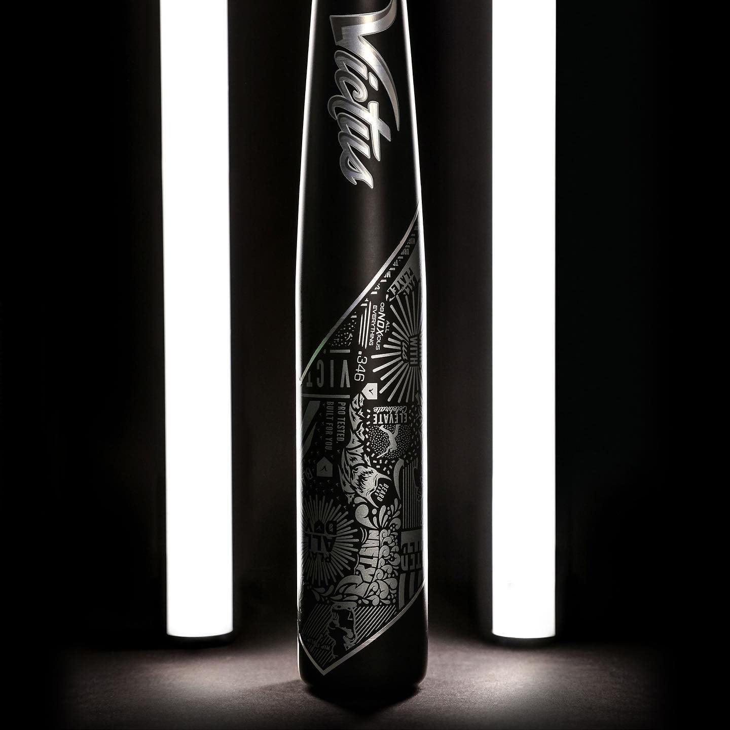 A black Victus baseball bat with intricate white graphic designs is positioned upright between two vertical, glowing white lights on a dark background.