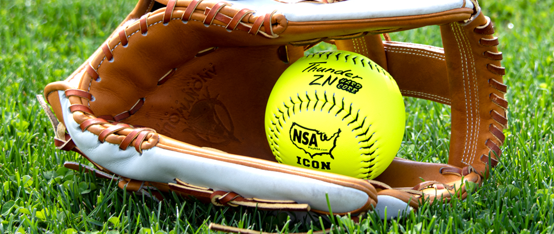 A yellow softball with black lettering rests inside an open brown leather glove on green grass.