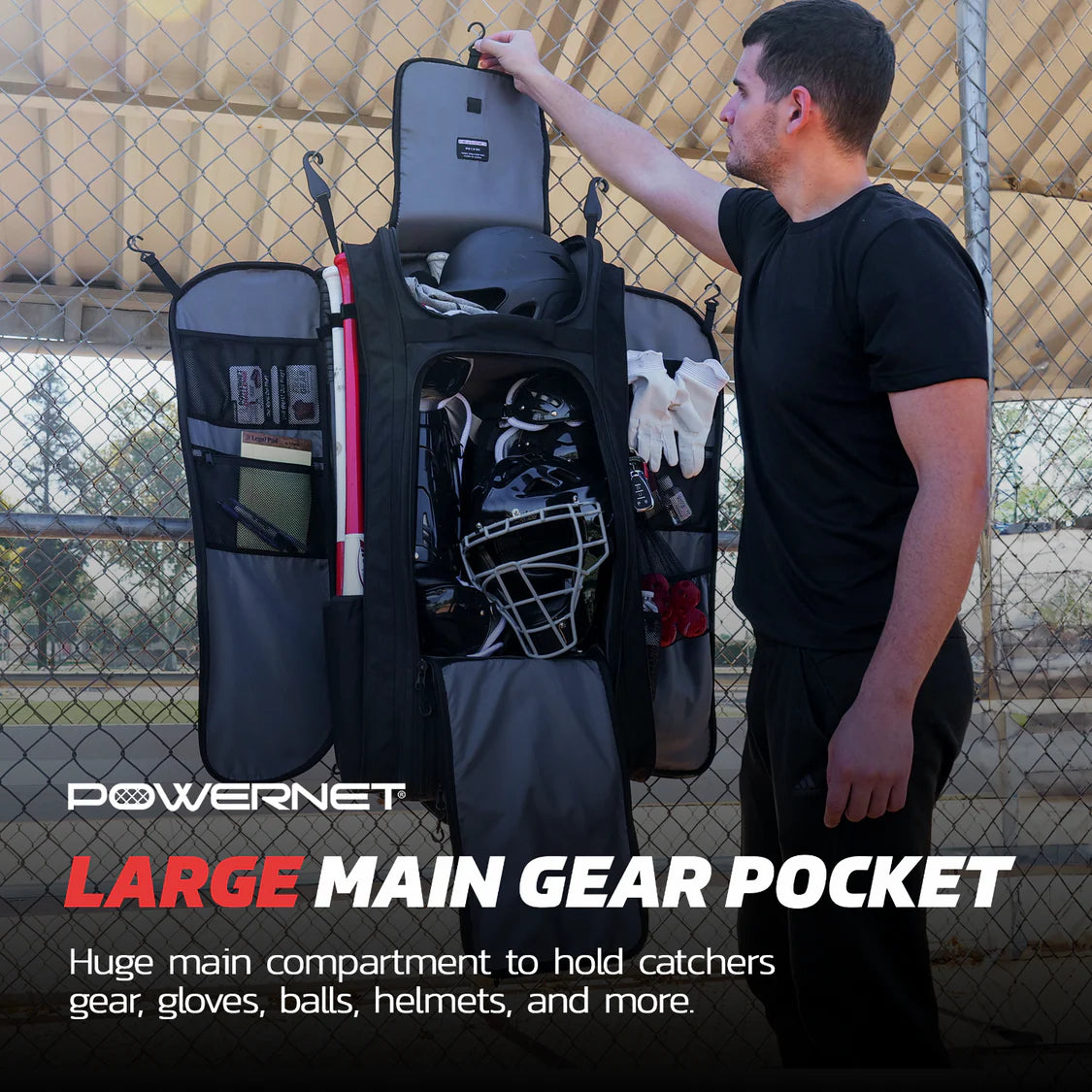 A man stands next to an open PowerNet Optimus Catcher's Gear Bag (B013-BK) hanging on a fence, showing a spacious compartment with catcher’s gear, gloves, balls, and a helmet. Text: PowerNet LARGE MAIN GEAR POCKET.