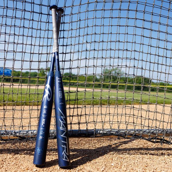 Two Rawlings 2025 Mantra (-10) Fastpitch Softball Bats (RFP4M10 DEMO) stand upright, leaning together on a dirt field by a net, with green grass and a clear blue sky—ideal for any Rawlings fastpitch bat enthusiast.