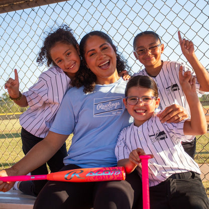 A smiling coach sits on a bench with three young girls in baseball uniforms, all celebrating. The girl in front proudly holds her 2026 Rawlings ALO78 (-12) Fastpitch Softball Bat (RFP6ALO12), with a field and fence behind them.