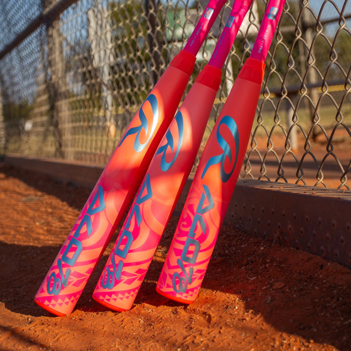 Three Rawlings ALO78 (-12) Fastpitch Softball Bats (RFP6ALO12) rest against a chain-link fence on a dirt field, with a blurred background of trees and a baseball diamond.