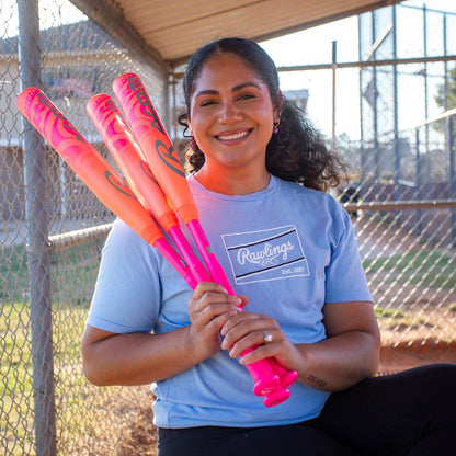 A woman with curly hair, in a light blue Rawlings t-shirt, smiles in a dugout while holding three bright pink and orange 2026 Rawlings ALO78 (-12) Fastpitch Softball Bats (RFP6ALO12). A chain-link fence and field are in the background.