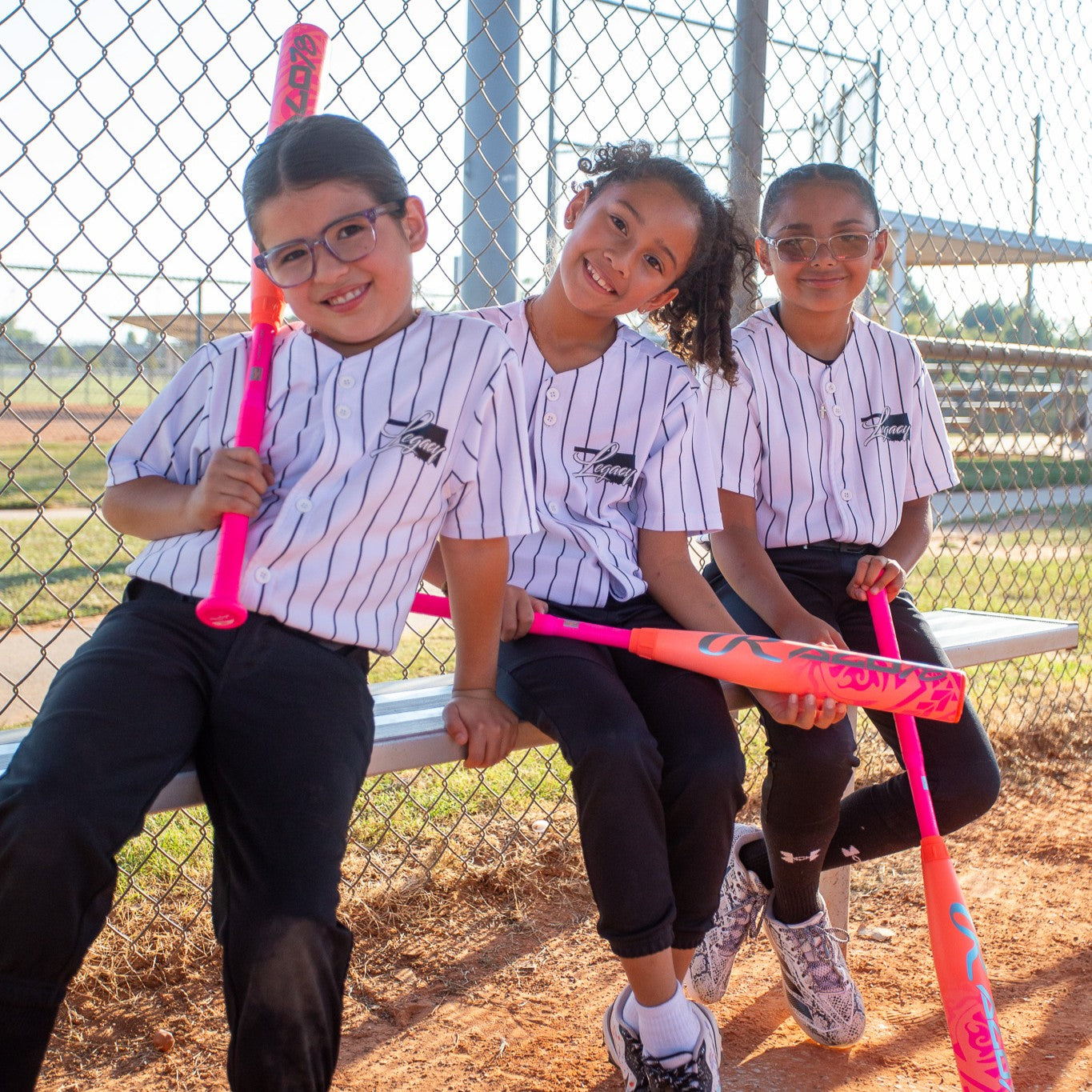 Three young girls in matching striped jerseys sit on a bench at a baseball field, smiling and holding colorful bats, including the Rawlings 2026 ALO78 (-12) Fastpitch Softball Bat (RFP6ALO12). Behind them: a chain-link fence and sunny sky.