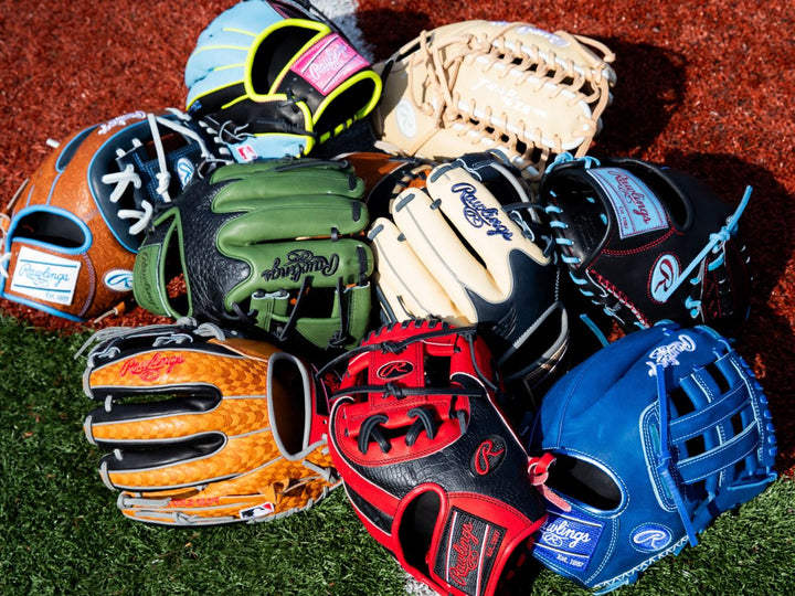 A pile of colorful baseball gloves, including blue, red, green, tan, and black Rawlings gloves, arranged on green artificial turf and reddish-brown infield dirt.