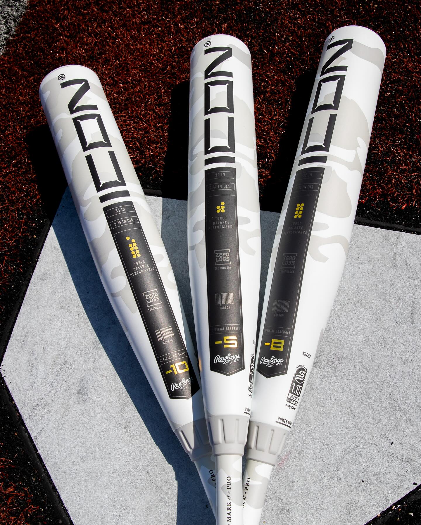 Three Rawlings 2025 Icon USSSA baseball bats with black, white, and gray accents lie on brown turf by a base, each with a unique drop: -10, -8, and the popular (-5) 2 3/4" RUT5I5 model.