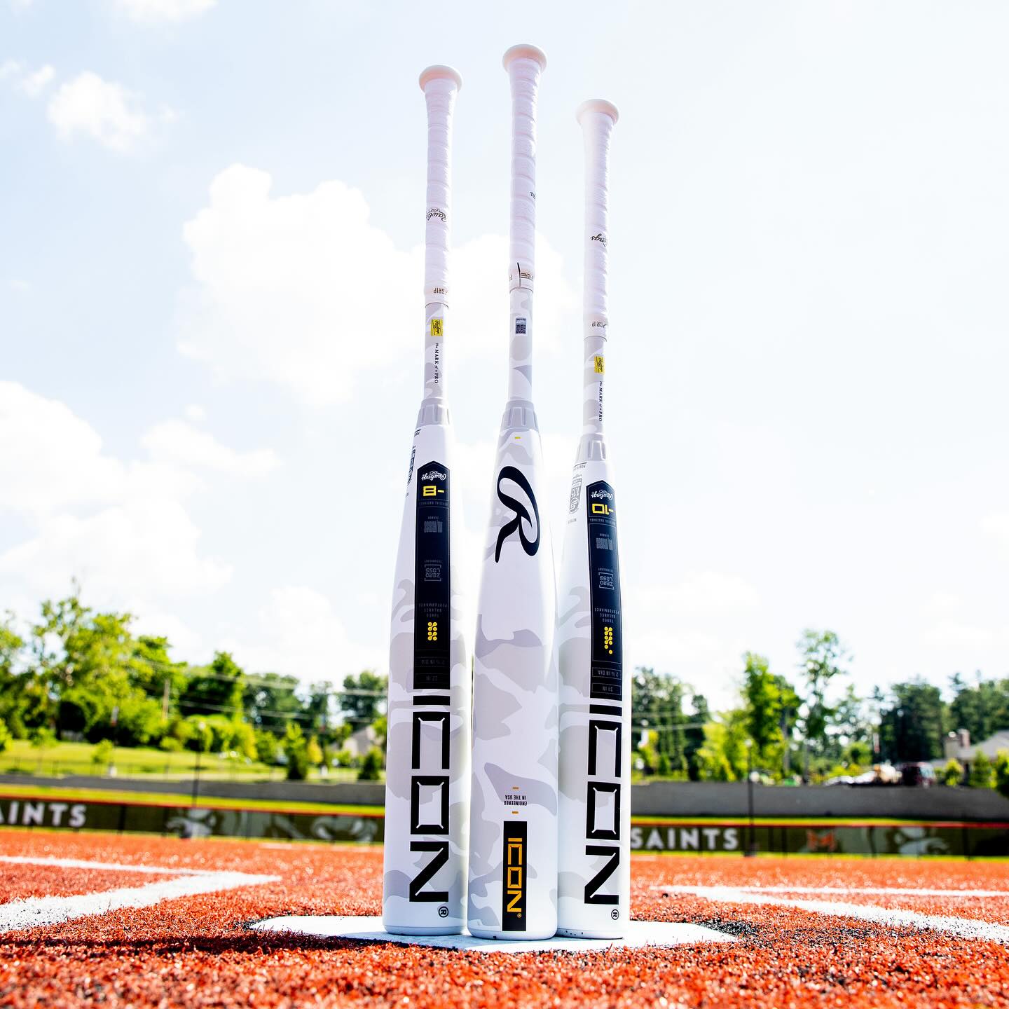 Three white 2025 Rawlings Icon (-5) 2 3/4" USSSA Baseball Bats (RUT5I5) by Rawlings stand upright on red turf, with green trees and a blue sky behind them. Each bat features a white grip and bold Rawlings branding.