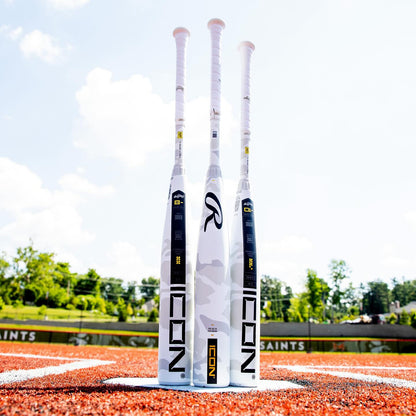 Three white 2025 Rawlings Icon (-5) 2 3/4" USSSA Baseball Bats (RUT5I5) by Rawlings stand upright on red turf, with green trees and a blue sky behind them. Each bat features a white grip and bold Rawlings branding.
