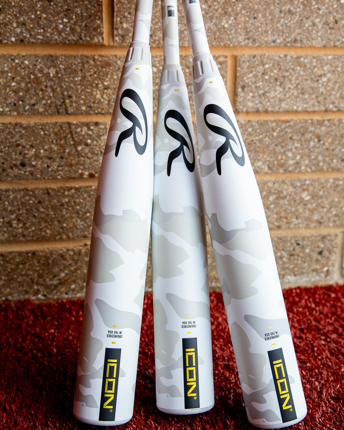 Three 2025 Rawlings Icon (-5) USSSA baseball bats with white carbon composite and light gray camo stand upright on red turf against a tan brick wall. Each bat features "ICON" in yellow and black lettering near the handle.