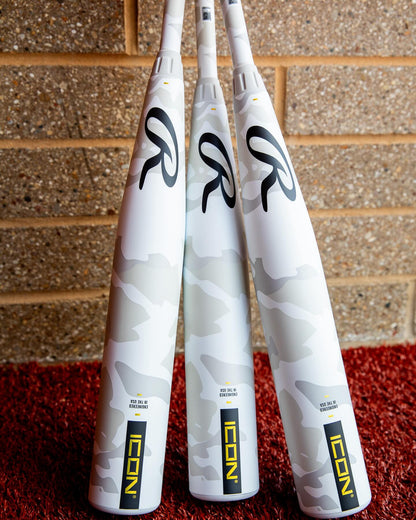 Three 2025 Rawlings Icon (-5) USSSA baseball bats with white carbon composite and light gray camo stand upright on red turf against a tan brick wall. Each bat features "ICON" in yellow and black lettering near the handle.