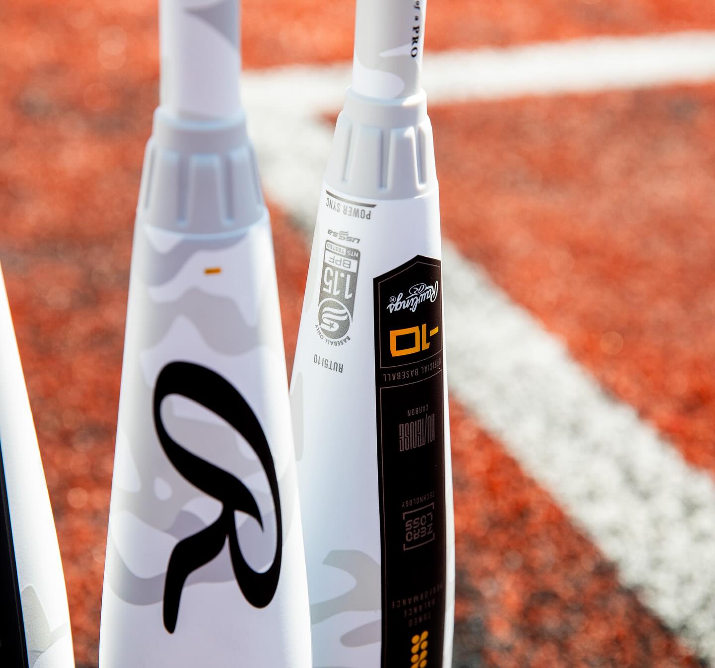 Close-up of the handles of three Rawlings 2025 Icon (-5) 2 3/4" USSSA Baseball Bats (RUT5I5), white with black and gold accents, standing upright on orange turf with a white line in the background.