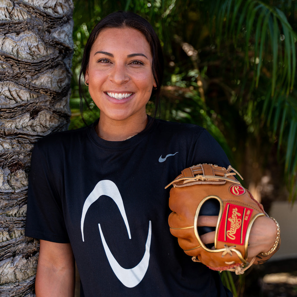 A smiling woman leans against a tree, wearing a black Nike shirt and holding a brown Rawlings baseball glove, with palm trees blurred in the background.
