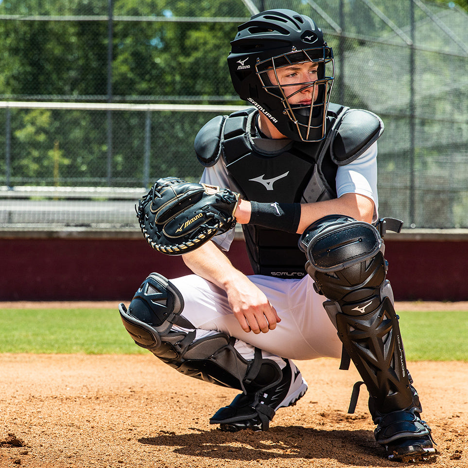 A baseball catcher wearing full protective gear crouches behind home plate on a sunny day, ready to catch a pitch. A fence and trees are visible in the background.