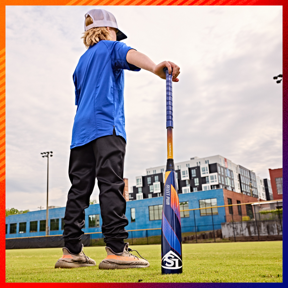A child wearing a blue shirt, black pants, and a backwards cap stands on a grassy field holding a baseball bat, with buildings and a cloudy sky in the background.