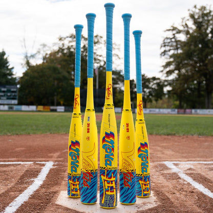 Five vibrant yellow and blue bats with blue grips stand upright at home plate, trees and a scoreboard behind them. Among these is the popular 2026 Victus Nova Lit (-8) USSSA Baseball Bat (VSBNL8) from Victus.