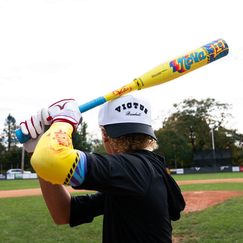 A baseball player in a white Victus cap and colorful gear holds a 2026 Victus Nova Lit (-8) 2 3/4" USSSA Baseball Bat (VSBNL8) over their shoulder on the field, with trees and a scoreboard visible in the background.