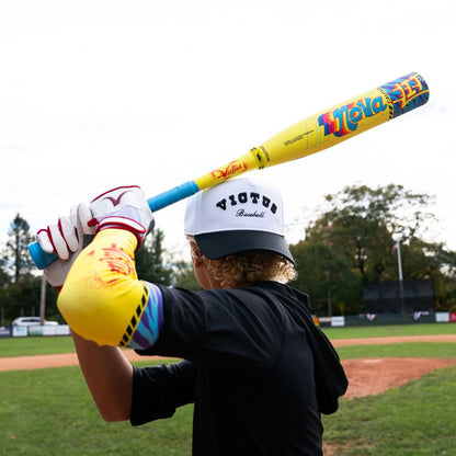 A baseball player in a white Victus cap and colorful gear holds a 2026 Victus Nova Lit (-8) 2 3/4" USSSA Baseball Bat (VSBNL8) over their shoulder on the field, with trees and a scoreboard visible in the background.