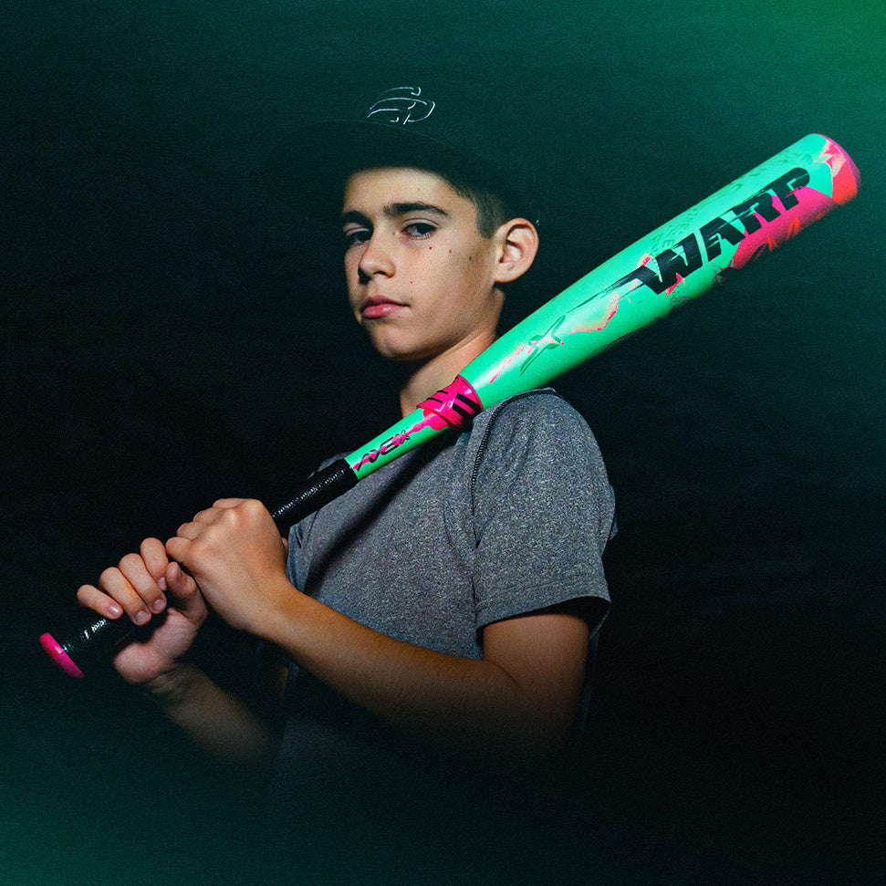 A young boy in a gray shirt and black cap holds a colorful neon green and pink baseball bat across his shoulders, looking confidently at the camera against a dark background.