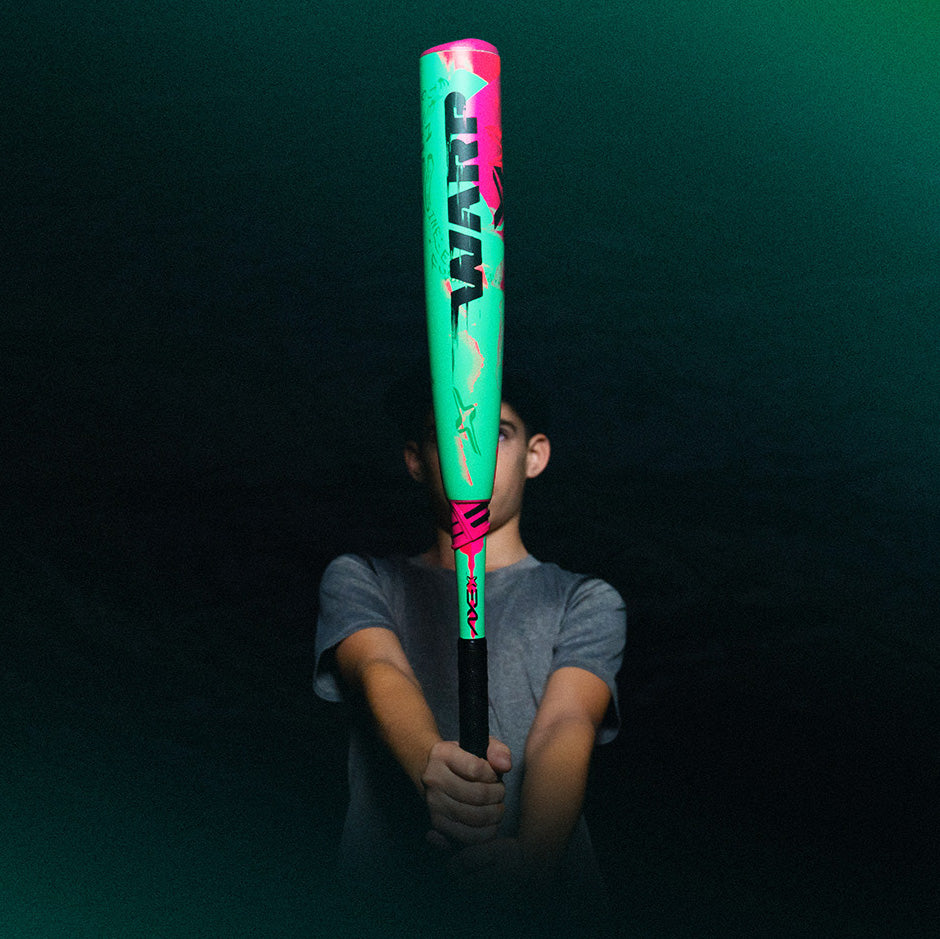 A person in a gray shirt holds a brightly colored neon baseball bat vertically in front of their face against a dark background.