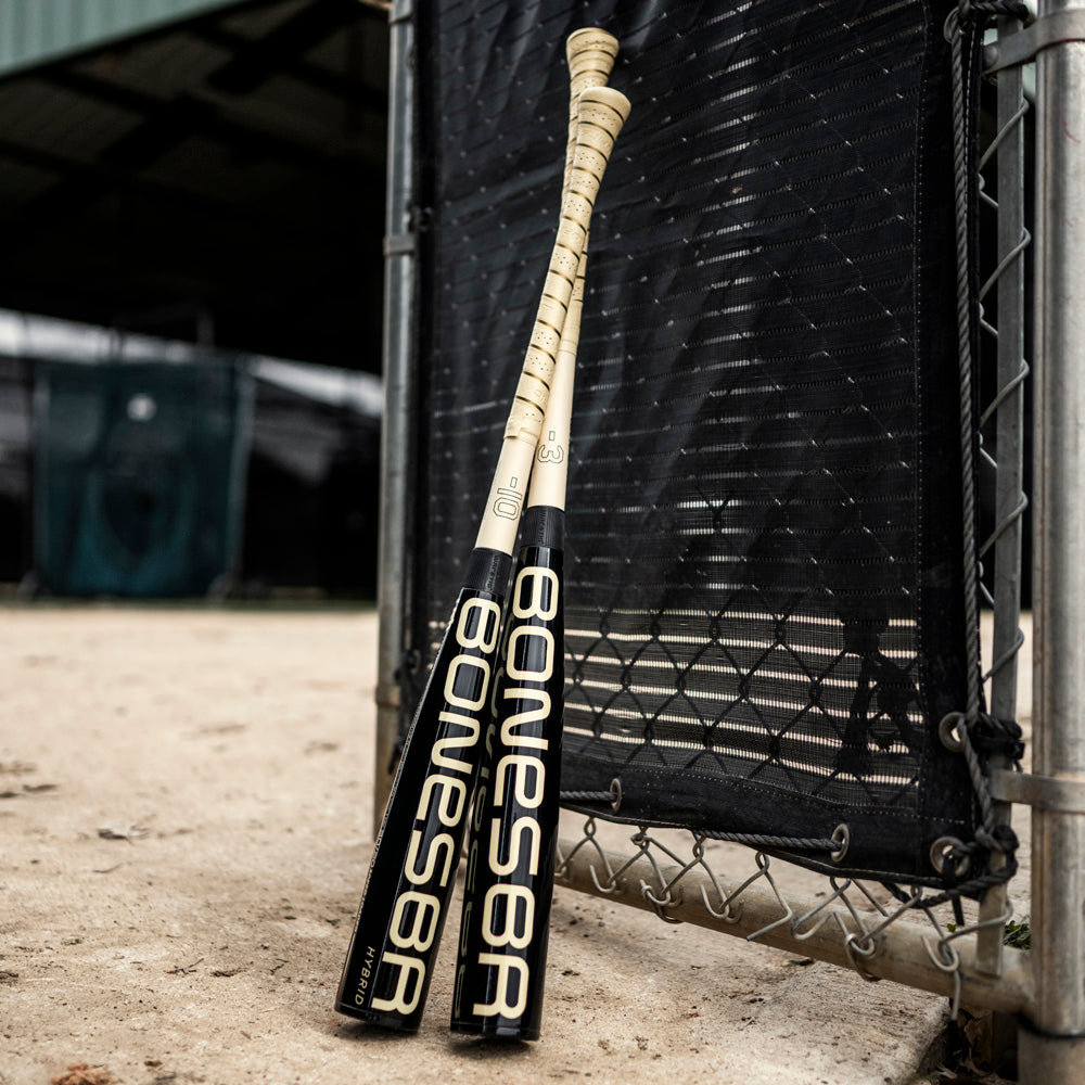 Two black and gold Warstic baseball bats, including the 2025 Bonesaber Hybrid Black Cobra (-3) BBCOR, stand upright against a black net fence on a baseball field, with dirt ground and a metal gate partially visible.