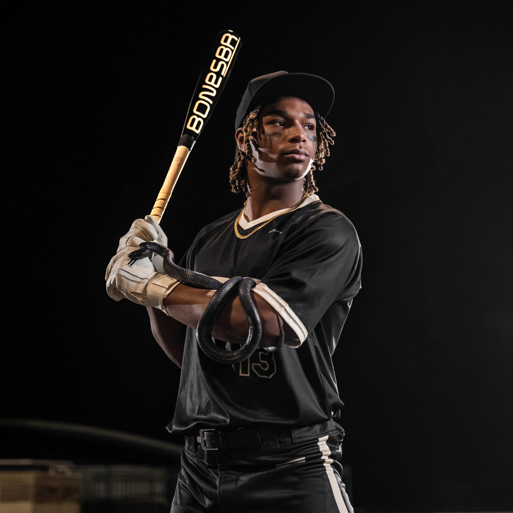 A focused baseball player in black grips a Warstic 2025 Bonesaber Hybrid Black Cobra (-3) BBCOR bat. The dark setting highlights his stance and the bold yellow BONESABA label on the bat.