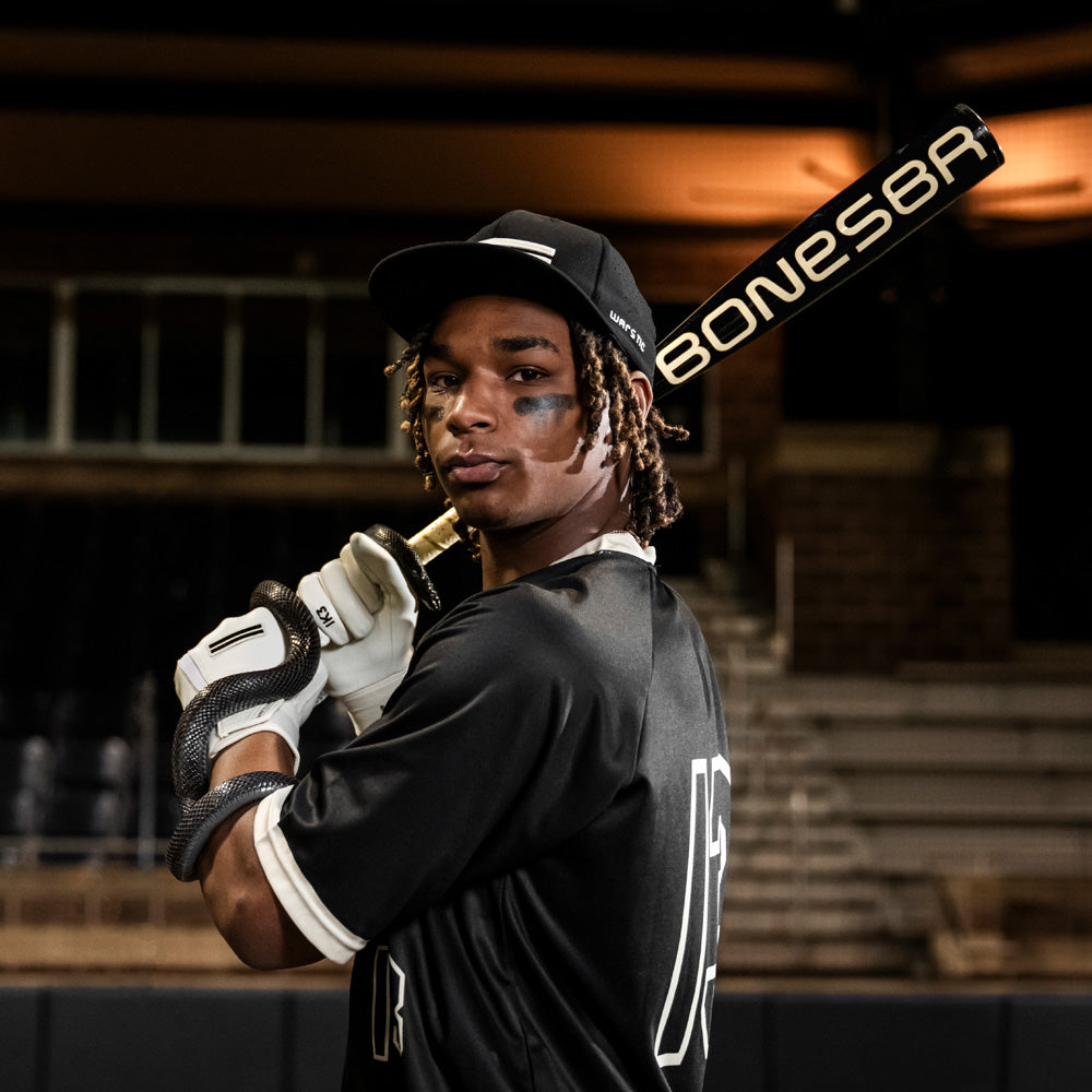 A baseball player in a black uniform and cap confidently poses with a Warstic 2025 Bonesaber Hybrid Black Cobra (-3) BBCOR Baseball Bat over his shoulder in a dimly lit stadium.