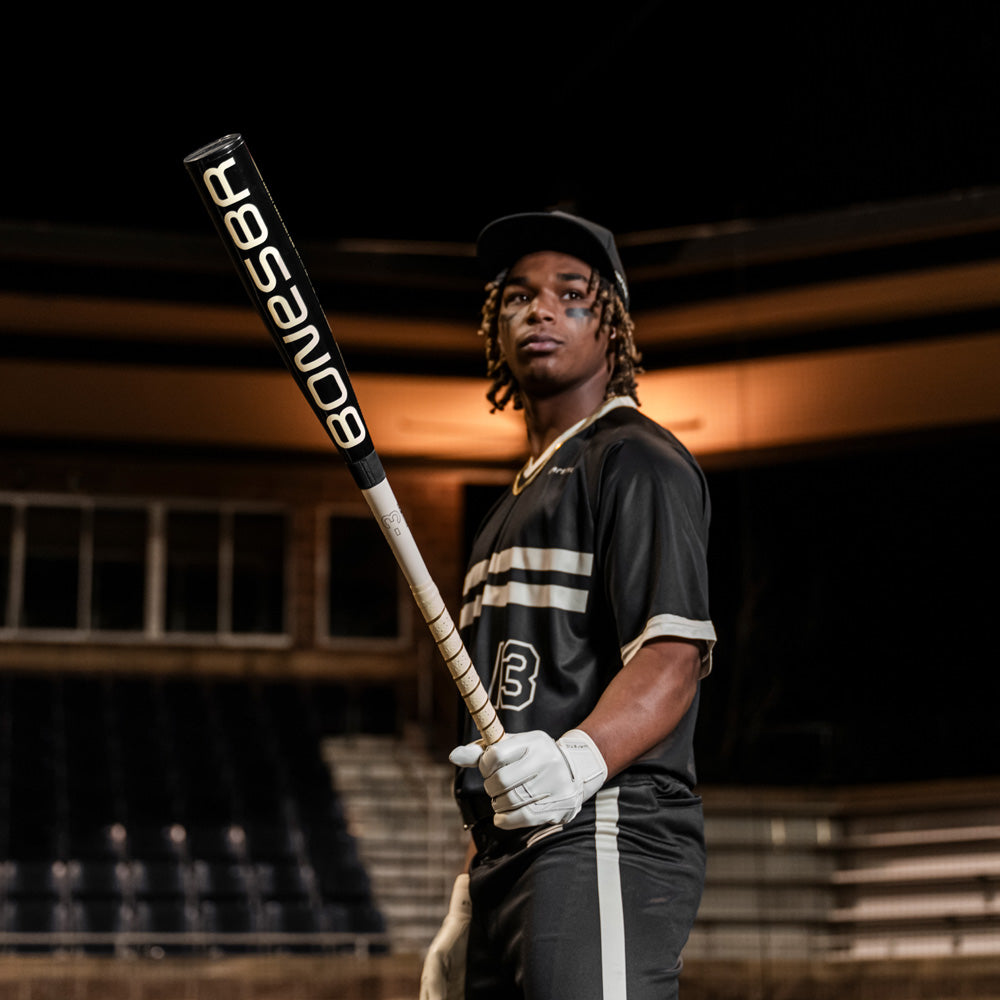 A baseball player in a black uniform with white stripes holds the 2025 Warstic Bonesaber Hybrid Black Cobra (-3) BBCOR Baseball Bat (MBBSRHB25BK3) in a stadium at night, wearing a cap and batting gloves.