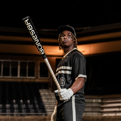 A baseball player in a black uniform with white stripes holds the 2025 Warstic Bonesaber Hybrid Black Cobra (-3) BBCOR Baseball Bat (MBBSRHB25BK3) in a stadium at night, wearing a cap and batting gloves.