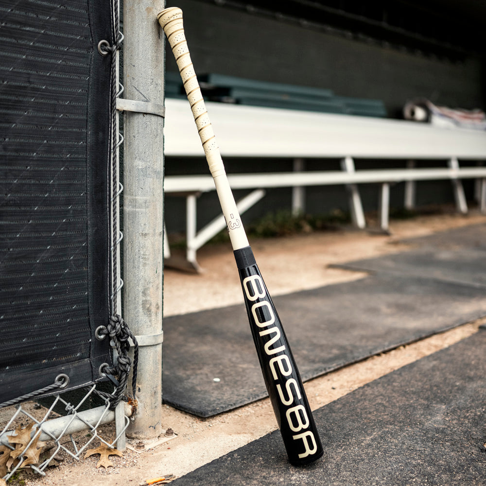 A 2025 Warstic Bonesaber Hybrid Black Cobra (-3) BBCOR Baseball Bat leans against a fence by a dugout with white benches and a dirt path.