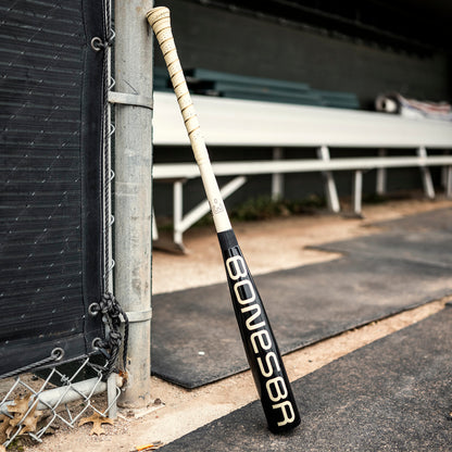 A 2025 Warstic Bonesaber Hybrid Black Cobra (-3) BBCOR Baseball Bat leans against a fence by a dugout with white benches and a dirt path.